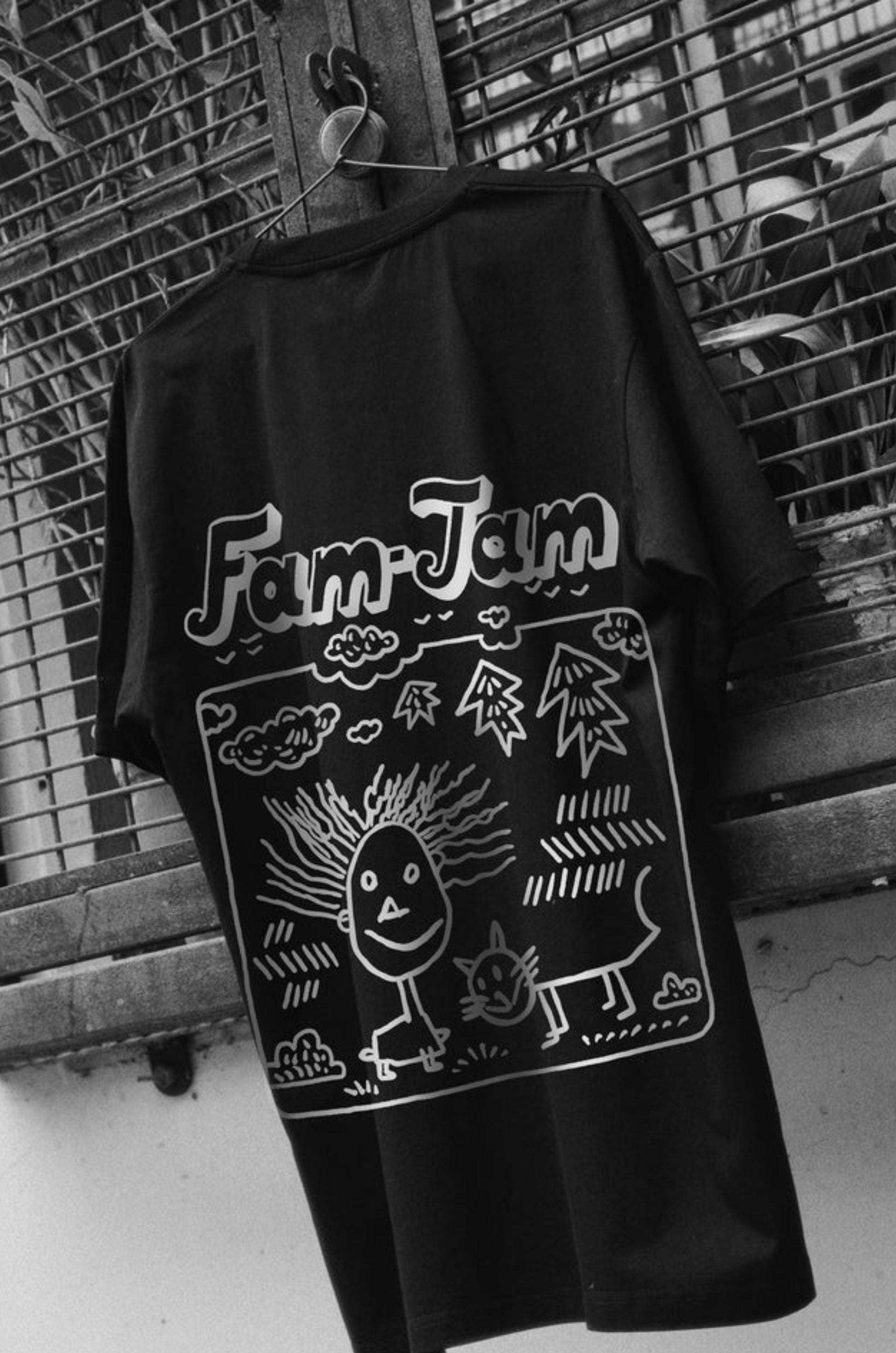 Oversized black T-shirt with a round neck and short sleeves, featuring a white print on the back with the text 'FAM-JAM' and various drawings, hung on a fence, by Blaknwhite