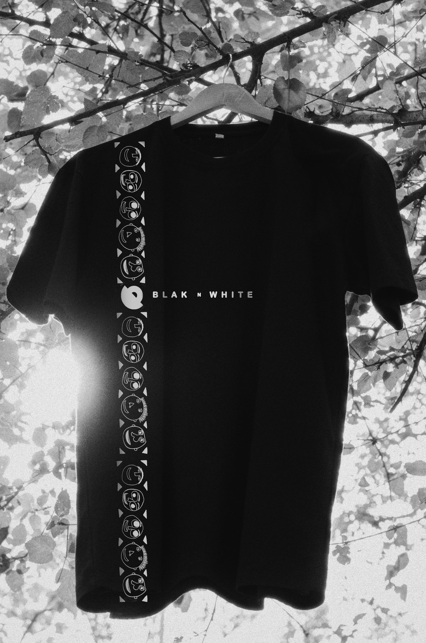 Oversized black t-shirt with short sleeves and round neckline, featuring a design with what appears to be floral patterns or moody illustrations along the front and sleeves, hung on a tree branch with a blurred background.